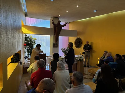 Chapel interior with priest at lectern, large crucifix, candles, congregation seated, yellow walls.