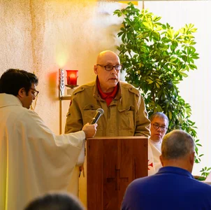 Older man in tan jacket speaks at wooden podium; someone holds microphone; candle, plant.