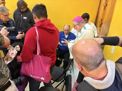 Bishop in white vestments and pink zucchetto blesses a man as photographers circle in a yellow room