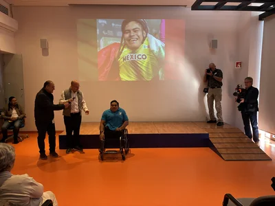 Wheelchair athlete interviewed; two men speak; wall projection shows smiling athlete in Mexico jersey