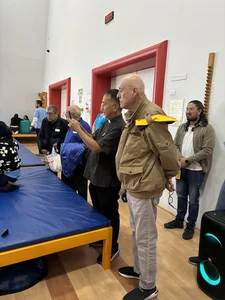 Group watches event in gym with blue mats; bald man in tan jacket with yellow epaulette; others.