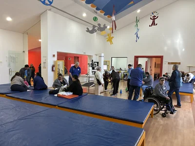Bright therapy room with blue mats; people and volunteers help; a man in a wheelchair sits at mats.