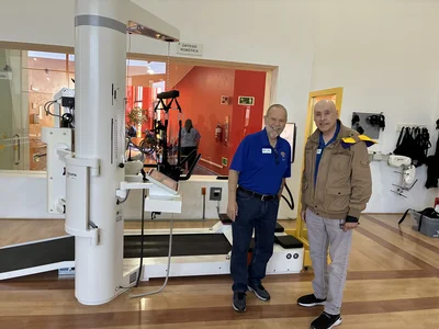 Two men in blue shirts smile beside a large white robotic rehabilitation device in a clinic.