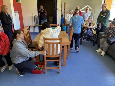 Group of seniors around a table as a volunteer kneels by a tub of colorful balls; photographer.