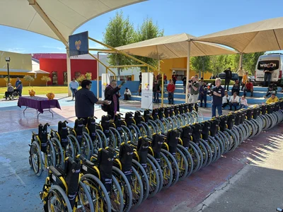 Row of yellow-framed wheelchairs at outdoor event; people, photographers, colorful buildings.