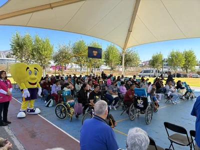 Outdoor event under a large canopy; yellow heart mascot near seated audience, many in wheelchairs.