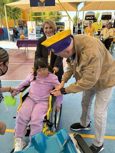 Girl in wheelchair with yellow frame guided by man wearing yellow-blue cap at Teleton charity event.