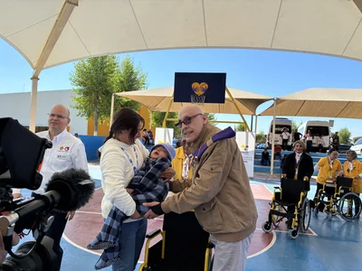Under a canopy at a charity event, woman hands baby to older man; wheelchairs and volunteers nearby.