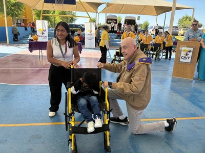 Older man kneels by a yellow wheelchair; child in it, woman stands smiling; Teletón event.