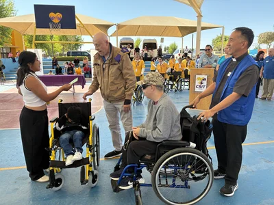 Outdoor basketball court event; woman hands card to boy in yellow-framed wheelchair; onlookers.