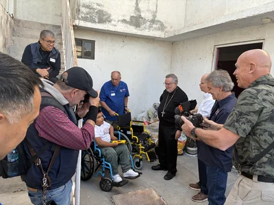 Media crew photographs a boy in a wheelchair; a priest and others observe outside a building.
