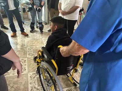 Caregiver in blue scrubs pushes a yellow wheelchair; a boy sits while onlookers photograph.