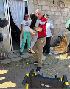 Man in red hugs child outside a stone house; onlookers nearby; Knights of Columbus wheelchair.