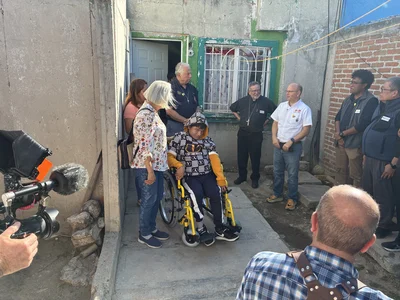 Media crew films as group gathers around a person in a wheelchair outside a worn building.