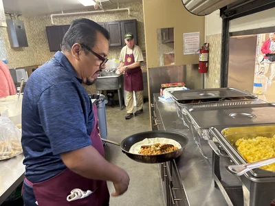 Cafeteria cook in blue shirt and maroon apron at skillet; eggs nearby; coworker at sink.