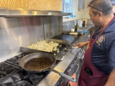 Male cook in maroon apron flips diced potatoes on a stainless griddle; skillet on front burner.