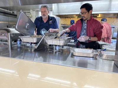 Two cooks in a stainless kitchen serve food from aluminum trays on a counter.
