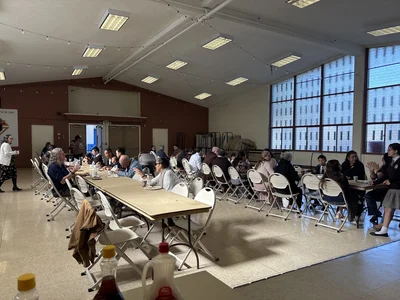 Community hall dinner: long tables, many diners, white folding chairs, string lights, windows.
