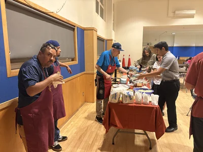 Volunteers in maroon aprons serve food at a buffet; people line up beside the table.