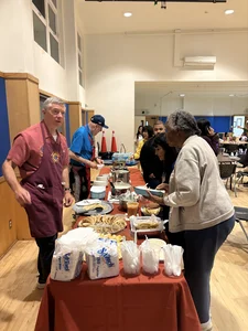 Community meal in a gym: volunteers in aprons serve food from red table; attendees queue.