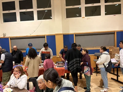 Diverse group at a community kitchen, serving and eating around a long table set with red cloth.