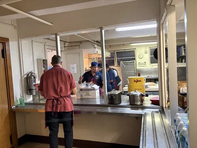 Pass-through kitchen: man in red shirt and apron at counter; others work in back; pots, igloo bucket.