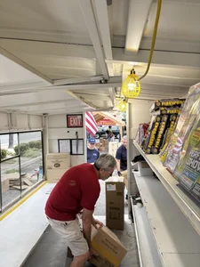 Man in a red shirt lifting a cardboard box inside a trailer with shelves of fireworks.