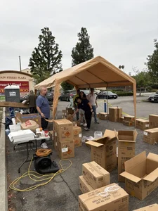 Volunteers organize many cardboard boxes under a tan canopy in a parking lot for distribution.