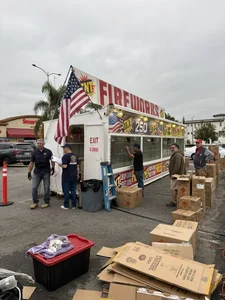 Workers unloading TNT boxes near a white fireworks trailer with American flags in a parking lot.
