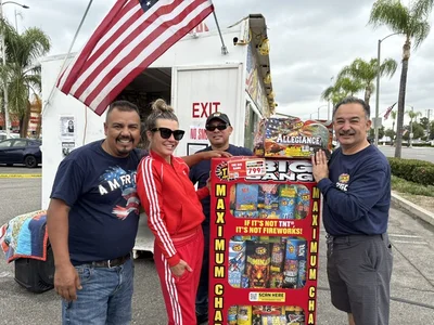 Four smiling people pose by a colorful fireworks display and American flag near a white truck.