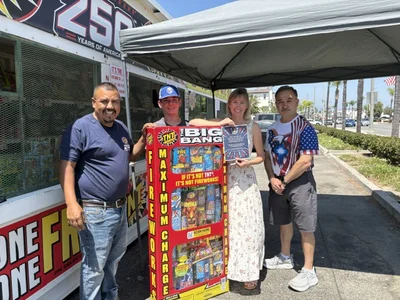 Four people pose with a large TNT fireworks display and certificate under a canopy by a truck.