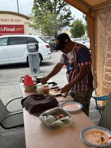 Man in an American flag shirt prepares food at a table under a canopy in a parking lot.