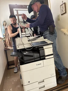 Two men, one in a hat and one in a tank top, work on a large white Canon printer indoors.