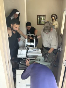 A nun and several men work together to move large printers through a narrow, cramped hallway.