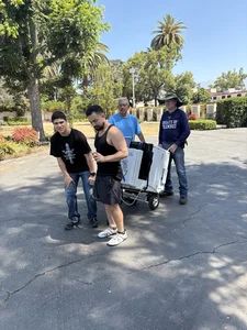 Four men push a large white equipment cart on wheels through a sunny, paved outdoor area.