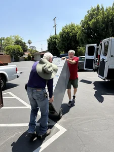 Two men tilt and move a large grey metal cabinet on wheels in a sunny parking lot.