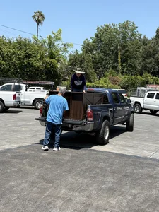 Man in blue shirt and woman in hat loading a large wooden crate into a dark blue pickup truck.