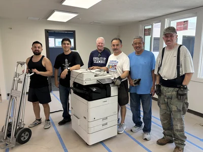 Six men pose in an office around a large white Canon printer, with a hand truck on the left.