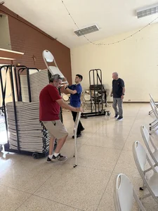 Three men move large stacks of white plastic chairs on a rolling cart in a spacious hall.
