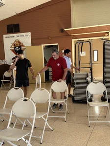 Three men arrange white folding chairs in a hall with a Knights of Columbus banner.
