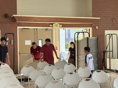 People move and stack white plastic chairs in a brick-walled room near an exit sign.