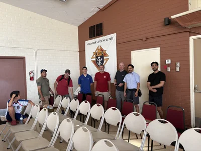 Seven men pose in a hall with a Knights of Columbus banner and rows of empty white chairs.