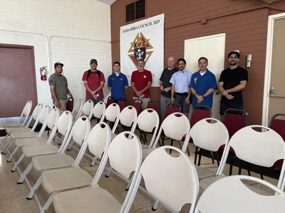 Eight men pose in a row in front of an Alhambra Council 2431 banner and rows of white chairs.