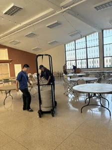 Man in a blue shirt pushes a cart of white plates through a large dining hall with round tables.