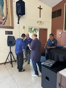 Four men in blue shirts setting up audio equipment near speakers and a mixer in a church hall.
