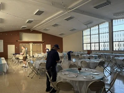 People preparing a bright, large banquet hall with round tables, white linens, and windows.